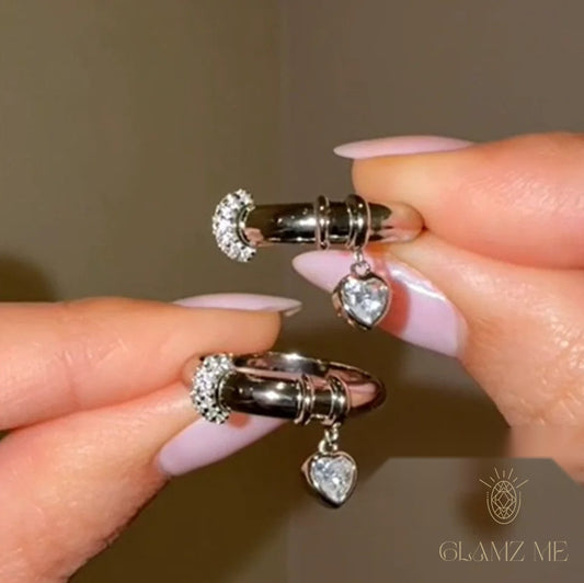 A close-up of a person's hands with light pink manicured nails holding a pair of silver huggie hoop earrings with heart-shaped crystal dangles, set against a neutral warm brown background with a 'GLAMZ ME' logo in the bottom right corner.