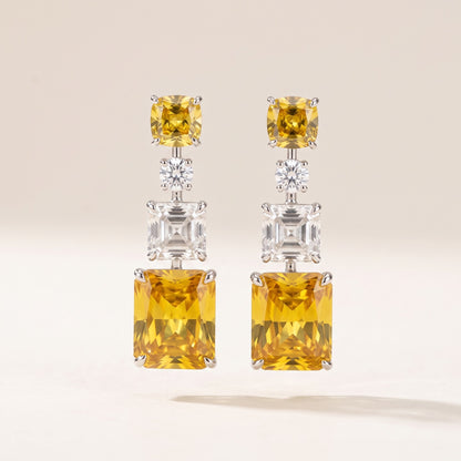 A pair of silver drop earrings featuring a large yellow cushion-cut gemstone at the top, a small clear round-cut stone in the middle, and a larger clear rectangular gemstone below, set against a warm off-white background.