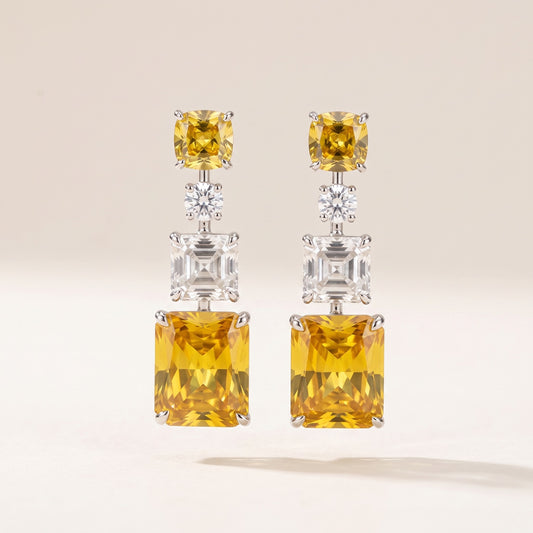 A pair of silver drop earrings featuring a large yellow cushion-cut gemstone at the top, a small clear round-cut stone in the middle, and a larger clear rectangular gemstone below, set against a warm off-white background.