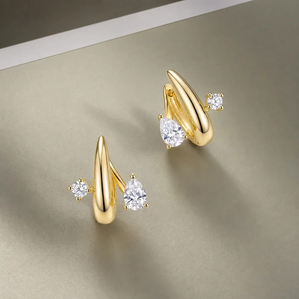 A pair of gold-toned earrings with curved, pointed posts and clear gemstones, displayed on a neutral beige surface.