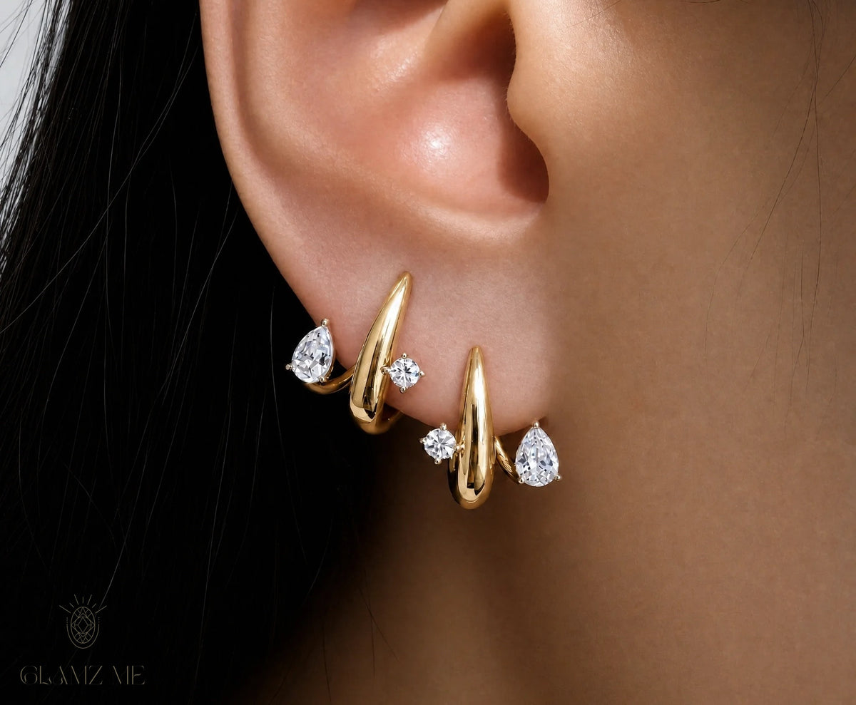 A close-up of a person's ear wearing a pair of gold huggie hoop earrings with small clear gemstones, on a dark background with a GLAMZAE watermark.