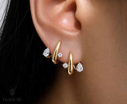 A close-up of a person's ear wearing a pair of gold huggie hoop earrings with small clear gemstones, on a dark background with a GLAMZAE watermark.