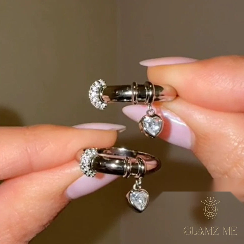 A close-up of a person's hands with light pink manicured nails holding a pair of silver huggie hoop earrings with heart-shaped crystal dangles, set against a neutral warm brown background with a 'GLAMZ ME' logo in the bottom right corner.