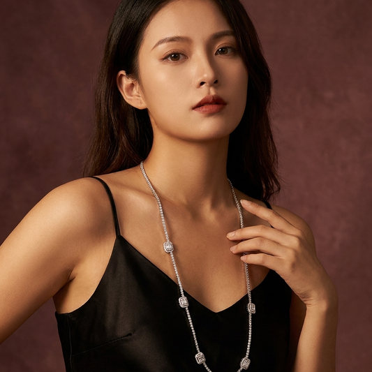 A woman wearing a long silver-toned necklace with square-cut clear stones, styled with a black satin camisole against a textured dark brown background.